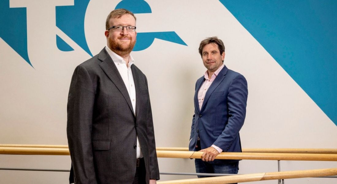 Two men standing separately from each other. One is holding on to a wooden handrail in the background.