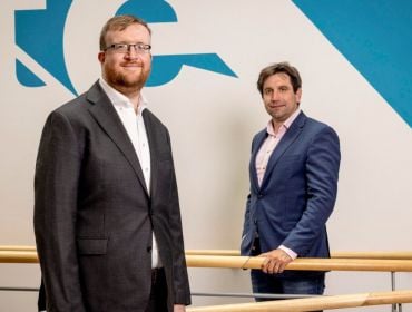 Two men standing separately from each other. One is holding on to a wooden handrail in the background.