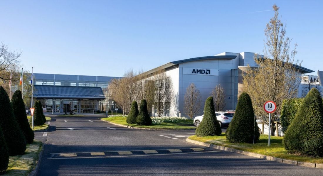 A large industrial building that is the AMD facility in Dublin. The front of the building and a large driveway with some trees and a blue sky are visible.