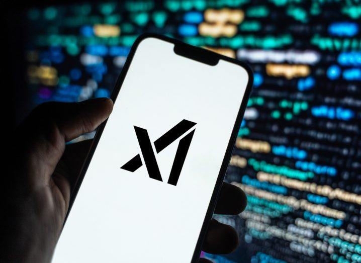 A person's hand holding a smartphone with the xAI logo on the screen,. A computer screen with code can be seen in the dark background.