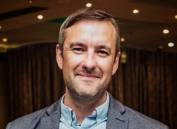 Headshot of DCU professor Andrew Kellett wearing a light blue shirt and a grey blazer inside what appears to be an auditorium.