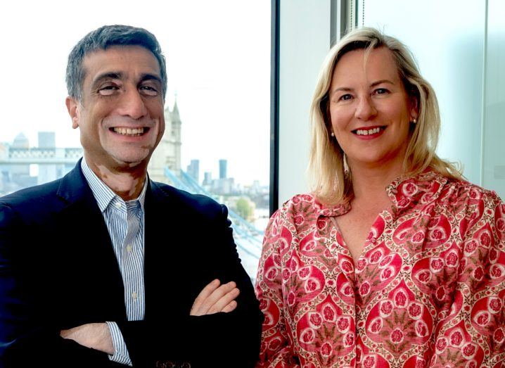 A man wearing a suit stands next to a woman in a red dress in what appears to be an indoor office space overlooking London.