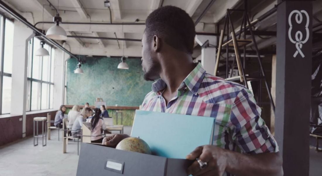 Young worker leaving an office carrying a box of belongings while he looks back at his colleagues in the distance.