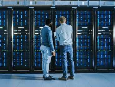 Two men standing facing a row of servers in a data centre.