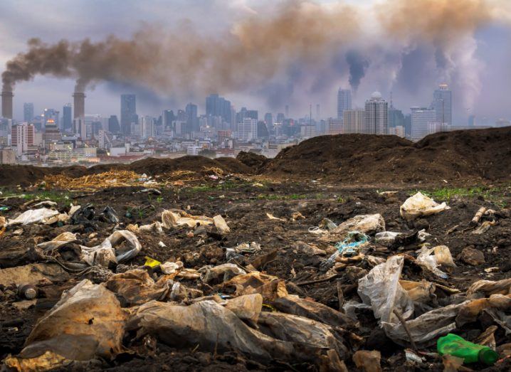 A wasteland with dirty rubbish bags and rotting rubbish with a polluted city and smoking chimneys in the background.