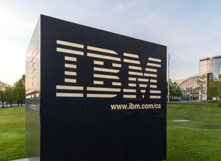 The IBM logo on a black square sign, with green grass and a building visible in the background.