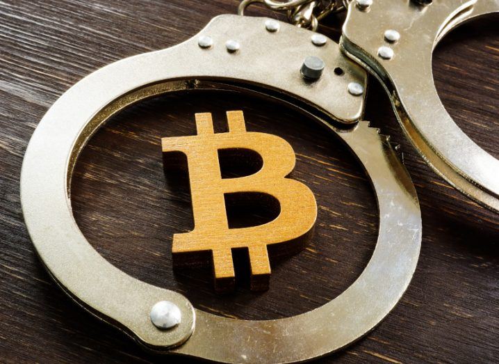 The Bitcoin logo on a wooden table, with a handcuff surrounding it. Used as a concept for crypto crime.
