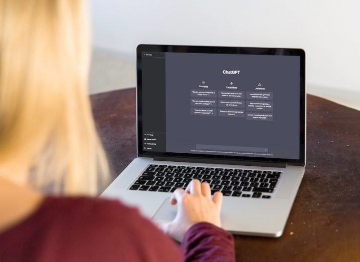 A person using a laptop that has the ChatGPT AI chatbot on the screen. The laptop is on a wooden table.