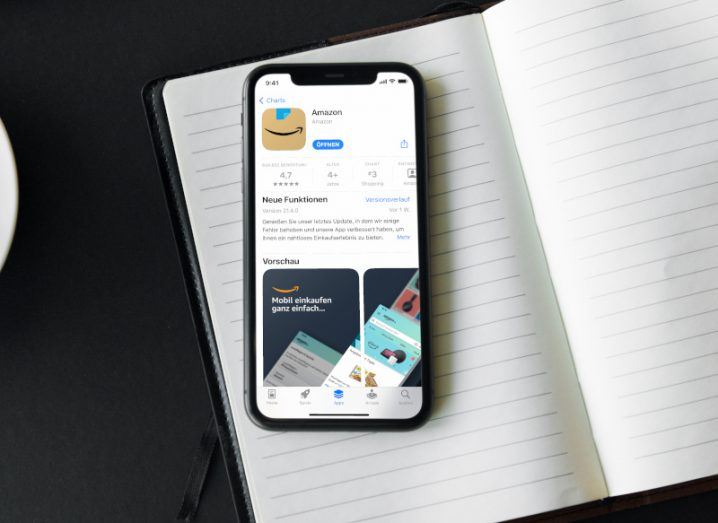 The Amazon store app page on a smartphone screen. The phone is laying on an open copy book, which is on a black surface.