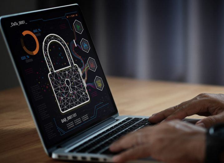 A person typing on a laptop that has a digital lock on the screen. The laptop is on a wooden table in a room. Used as a concept for cyberattacks and cybersecurity.