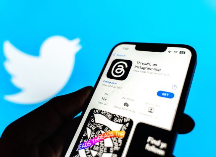 A phone displaying the Threads app on an app store being held by someone's hand. The Twitter blue bird logo is featured in the background behind the phone.