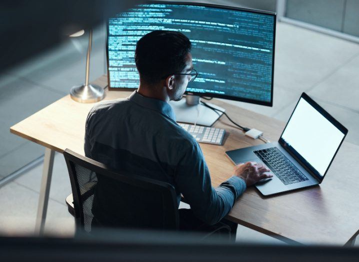A person sitting at a desk, typing on a laptop with a larger computer screen next to him.