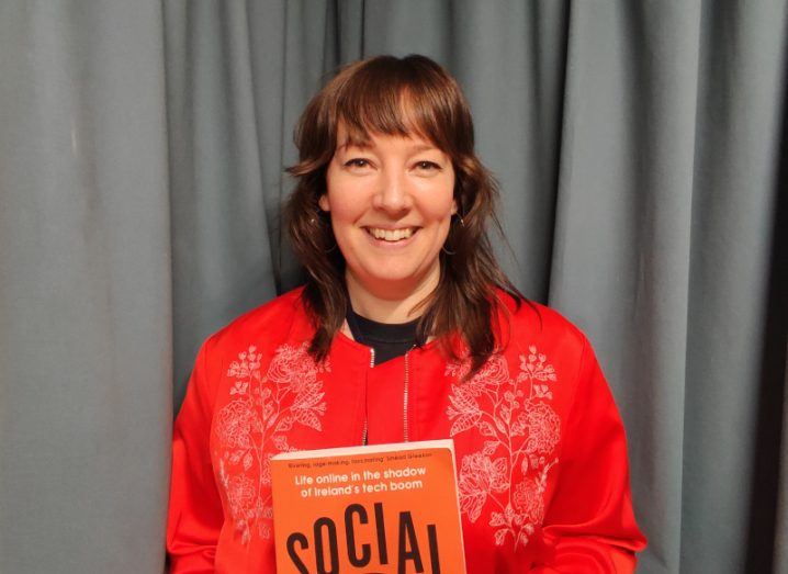 A woman stands against a grey curtain holding an orange book. It is journalist Aoife Barry.