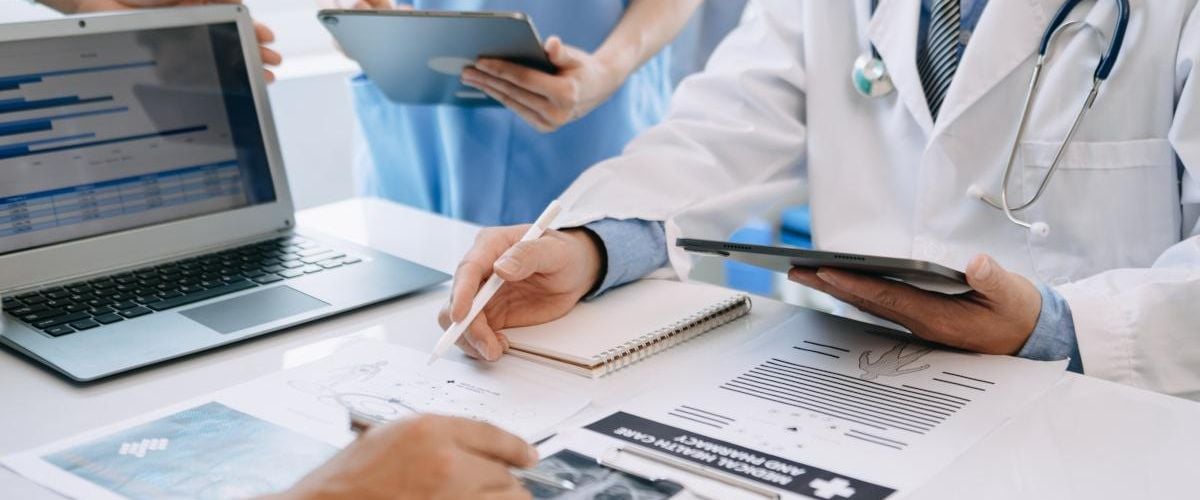 A group of medical staff stand around a desk full of medical records and documents, as well as a laptop.