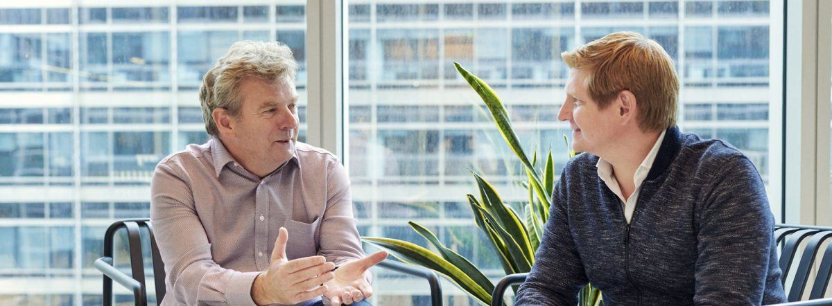 Two men sitting together and speaking, with a plant and a window in the background.