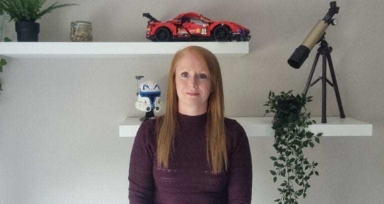 A woman wearing a maroon jumper stands smiling with a wall behind her. On the wall, there are two shelves with toys and plants on them. She is Lynn D'Arcy, a data centre engineer at Yahoo.