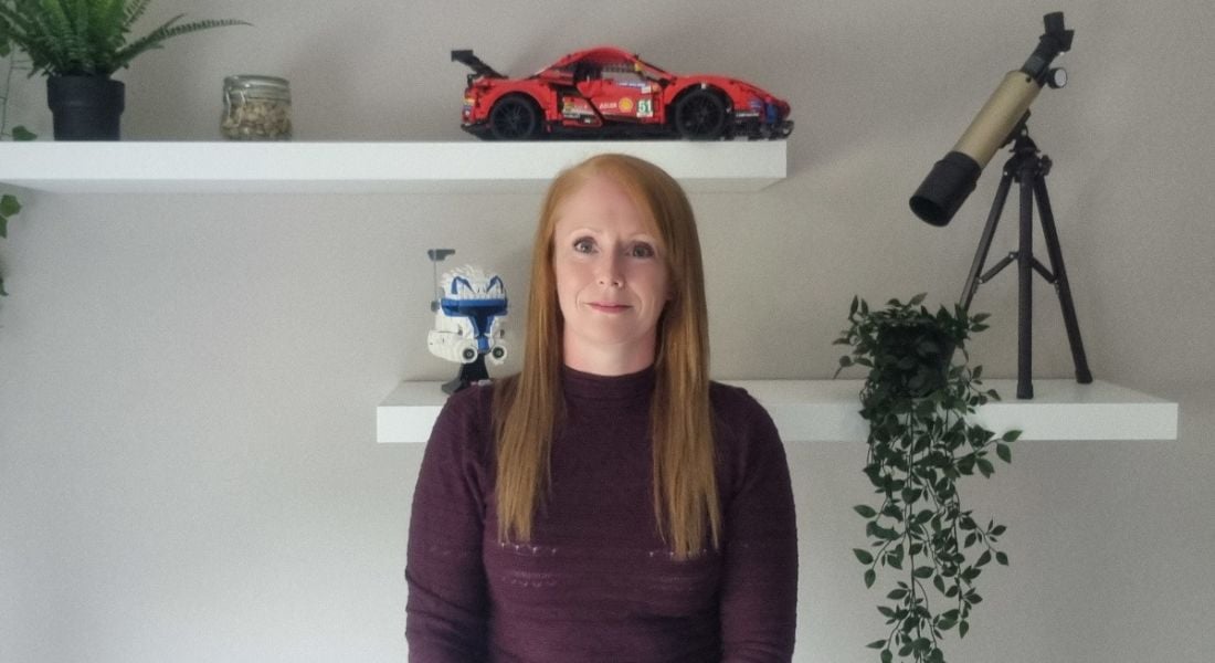 A woman wearing a maroon jumper stands smiling with a wall behind her. On the wall, there are two shelves with toys and plants on them. She is Lynn D'Arcy, a data centre engineer at Yahoo.