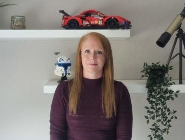 A woman wearing a maroon jumper stands smiling with a wall behind her. On the wall, there are two shelves with toys and plants on them. She is Lynn D'Arcy, a data centre engineer at Yahoo.