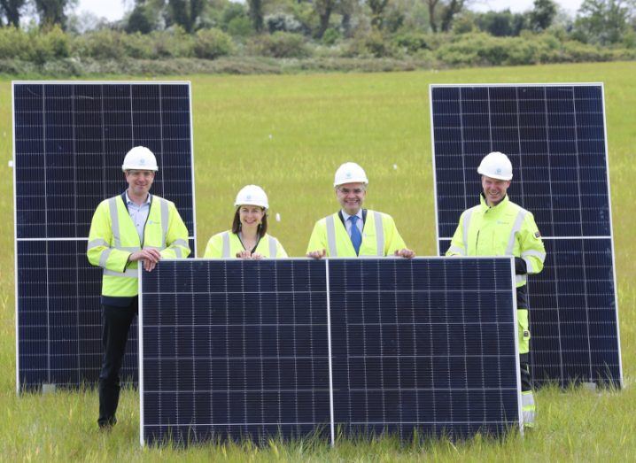 Three men and a woman wearing high visibility jacket and white hard hats, standing in a field behind a solar panel, with two upright solar panels behind them.