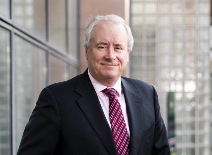 A man in a black suit and a red tie, with a building wall and windows behind him. He is Philip Maguire of Auxilion.