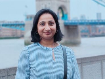 A woman smiles while standing with London's Tower Bridge sits behind her. She is Priyanka Lingegowda, a principal software engineer at Liberty IT.