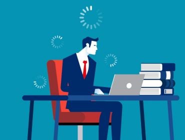 A man sits at a desk with a pile of books and a laptop. There are loading symbols floating around his head suggesting he’s not getting work done.