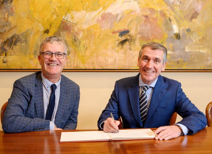 Two men sitting at a wooden table, with one man holding a pen to a sheet of paper. A painting is on the wall in the background.