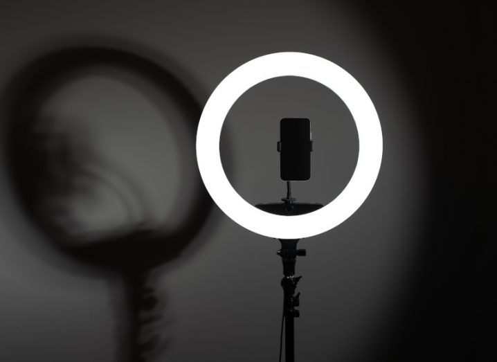 A ring light turned on in a dark room with a phone in the centre casting a shadow behind it.