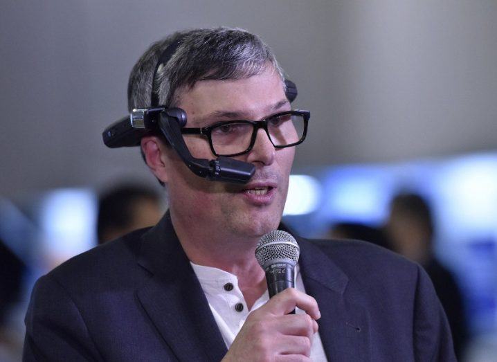 A man in a blue suit, holding a microphone and speaking. He is Simon Bain of OmniIndex.