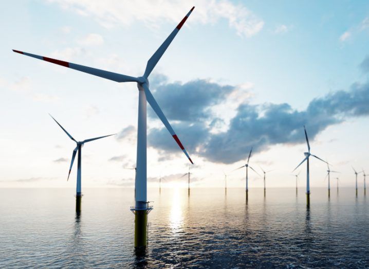 A group of wind turbines sit spinning in a large body of water with clouds and sun behind them.