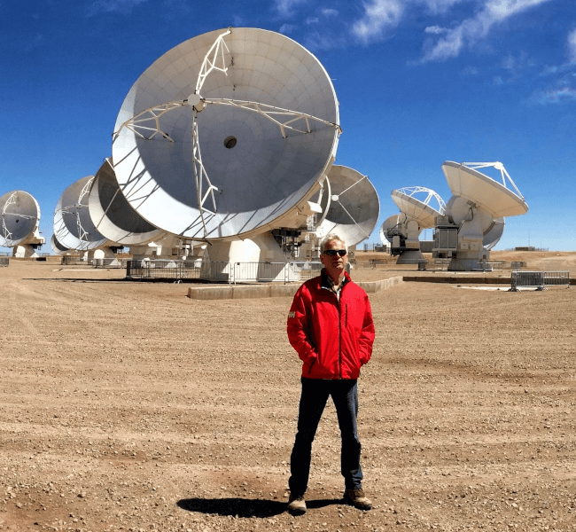 Prof Tom Ray standing in front of the ALMA space telescope in the Atacama Desert, Chile. There is a blue sky above and large radio telescope dishes in the background.